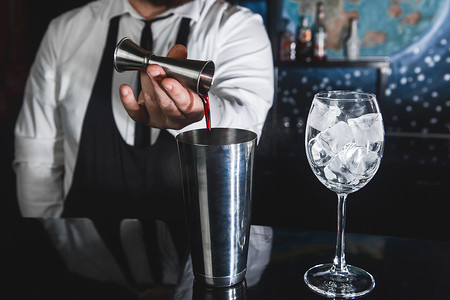 专业调酒师的手将糖浆倒入金属工具中的量杯中，用于制备和搅拌摇酒饮料的酒精鸡尾酒，以及装有冰块的玻璃杯