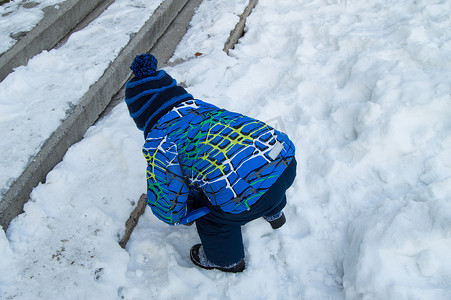 冬天，一个戴着蓝色针织帽和一件温暖夹克的小男孩在覆盖着雪的楼梯附近玩耍，从后面看