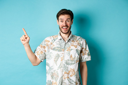 惊讶的快乐游客用手指指着空旷的地方，展示酷炫的促销，站在蓝色背景的夏威夷衬衫