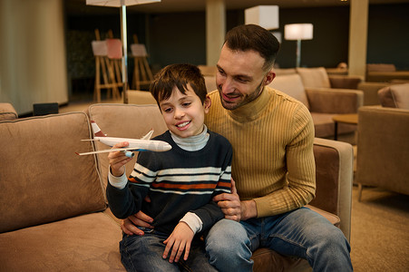 可爱的男孩玩玩具飞机，而他年轻慈爱的父亲坐在他旁边，在国际机场出发航站楼贵宾室等待登机时拥抱他