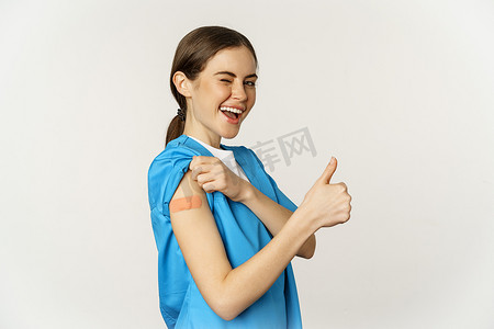 微笑的护士，穿手术服的医生，医务人员展示她接种疫苗的肩膀，竖起大拇指，建议接种冠状病毒疫苗，白色背景