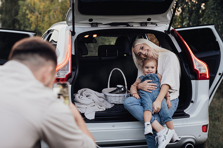 拍摄父亲摄影照片_丈夫拍摄他的妻子和蹒跚学步的女儿，他们坐在一辆 SUV 汽车的打开后备箱里，一家人在公路旅行度假