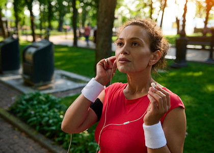 活跃的西班牙裔女性、女运动员、跑步者、戴着白色毛圈腕带的运动员，戴上耳机，清晨沿着城市公园慢跑时听音乐