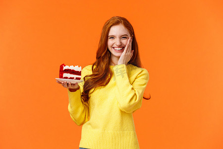 红发摄影照片_快乐可爱的白人红发女性拿着美味的奶油蛋糕，触摸脸颊，祝贺自己成为生日女孩，与家人庆祝生日，站在橙色背景