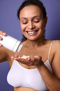 身穿白色内衣、微笑、开朗的非洲裔美国女性拿着一瓶食品补充剂，并将维生素复合物倒入手中。