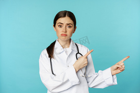 遗憾的表情摄影照片_失望的医生、女医务人员右手指着、看上去悲伤、遗憾、阴沉的表情、蓝色背景的肖像