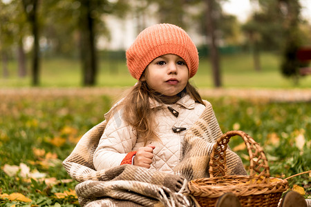 肖像小学龄前小女孩在橙色贝雷帽在黄色落叶在篮子里裹着毯子梦幻般地看着别处。