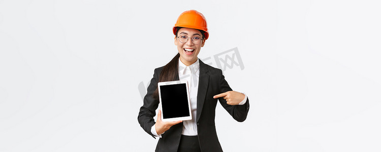 微笑的专业女性建筑经理、安全头盔和西装展示项目的亚洲工程师，用满意的表情、白色背景指着数字平板电脑屏幕