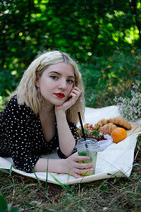 漂亮的金发女孩在公园里穿着白色格子野餐，拿着一个塑料透明杯和柠檬水。