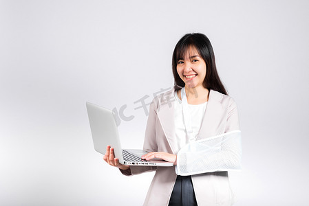 亚洲女商人在事故后自信地微笑着断臂，戴着手臂夹板接受治疗，但仍然需要工作，她拿着笔记本电脑