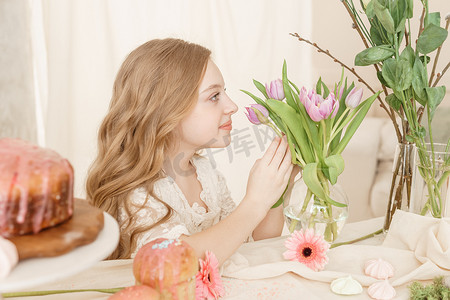 长发连衣裙女孩摄影照片_一个穿着浅色连衣裙、长发的女孩坐在复活节餐桌旁，桌上摆着蛋糕、春天的花朵和鹌鹑蛋。