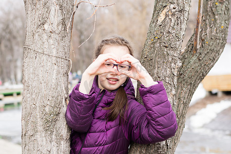 手指公摄影照片_早春，在城市公园里，戴眼镜的年轻漂亮女孩用手掌在脸前做出心形