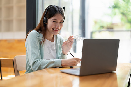 微笑的女学生佩戴无线耳机与老师在线学习，快乐的年轻亚洲女性学习语言听讲座观看网络研讨会写笔记看笔记本电脑坐在咖啡馆，远程教育