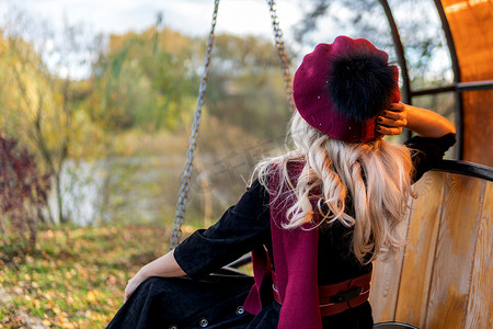 秋天，在池塘蓝云的映衬下，一个女人背坐在花园的秋千上，穿着酒红色外套，戴着双顶礼帽，眼睛美丽。