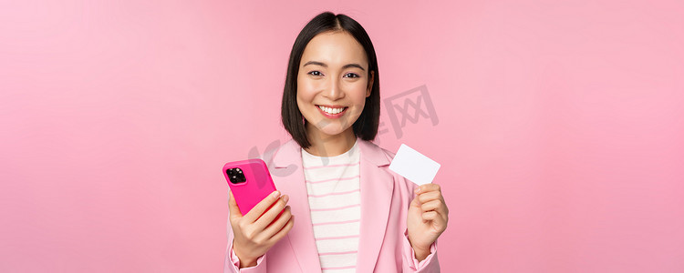 微笑快乐的亚洲女商人展示信用卡、在智能手机应用程序上在线支付、用手机订购、站在粉红色背景下的图片