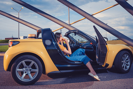 穿着蓝色牛仔裤、橙色上衣和人字拖的小女孩坐在黄色汽车敞篷车里吃汉堡。