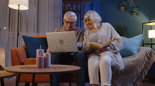 夜间客厅摄影照片_老祖父母夫妇读书，在家里夜间客厅的沙发上使用笔记本电脑