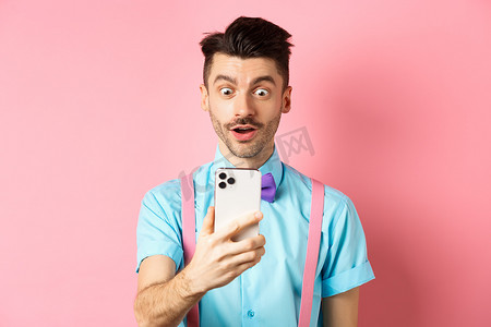 令人印象深刻的男人带着惊讶的表情阅读手机屏幕，看到在线促销优惠，站在粉红色的背景上