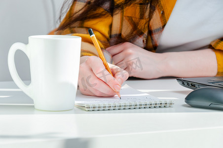 的铅笔摄影照片_特写镜头妇女手拿铅笔，在工作场所的记事本上写字，配有咖啡杯和笔记本电脑。