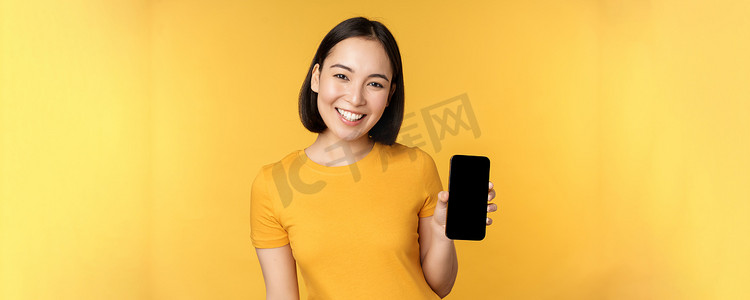 美丽快乐的亚洲女孩展示手机屏幕、智能手机小工具上的应用程序、站在黄色背景上