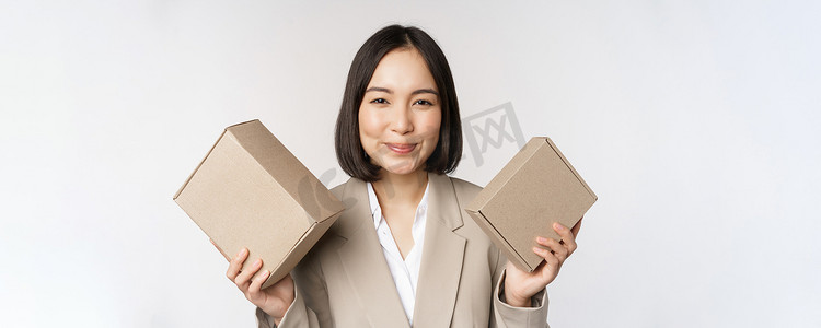 女脸手摄影照片_女售货员、亚洲女商人拿着装有公司品牌产品的盒子，对着镜头微笑，站在白色背景下的照片