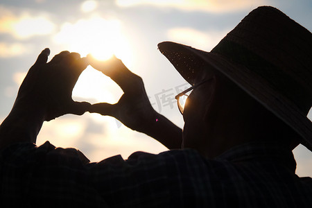 戴着帽子的高级农民的剪影，在日落时眺望远方，在金色的天空背景上形成心形。