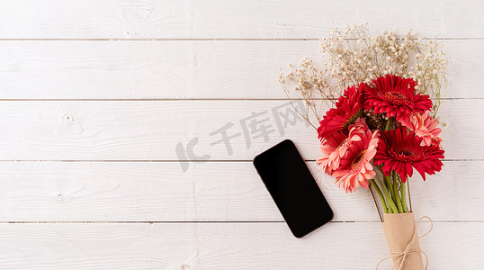 红色花卉邀请函摄影照片_白色木桌上的红色大丁草雏菊花和手机