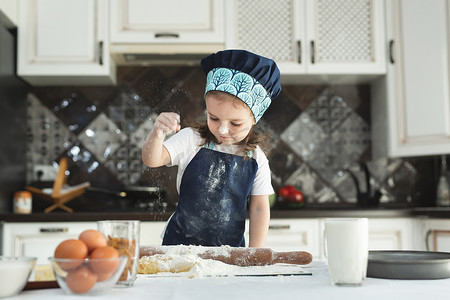 一个穿着围裙和厨师帽的小女孩在厨房里在面团上撒上面粉