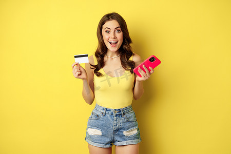 时尚的黑发年轻女性，拿着信用卡和智能手机，在商店里付款、购买或订购东西，站在黄色背景上