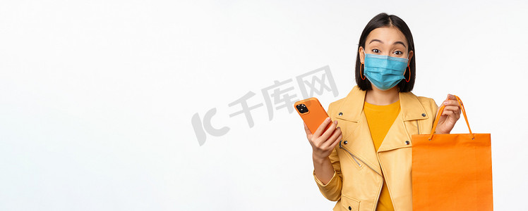 手机和app摄影照片_时尚的亚洲女孩购物者的形象，手持智能手机和没有商店标志的购物袋，戴着covid-19的医用面罩，白色背景
