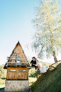 树木房子人摄影照片_新娘和新郎坐在一座木桥上，旁边是一座三角形的房子