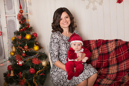 家庭环境摄影照片_妈妈和儿子穿着圣诞老人的服装，在靠近圣诞树的家庭环境中，靠着白色的木墙。