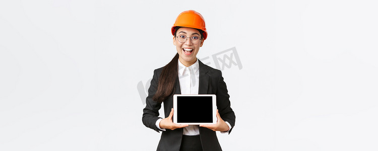 兴奋而惊讶的微笑亚洲女建筑工程师、身穿安全帽和西装的建筑师，展示数字平板电脑屏幕，表情惊讶，站在白色背景