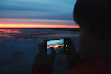 男子手持手机拍摄海上日落天空的照片。