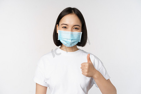 戴着医用面具的微笑的亚洲女孩竖起大拇指，认可一些好东西，赞扬和恭维公司，站在白色背景上