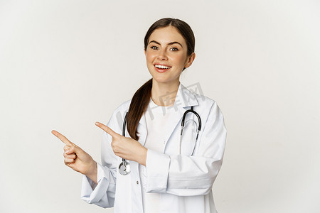 微笑的年轻女医生、医疗保健医务人员的肖像，手指向左，展示诊所促销、标志或横幅，站在白色背景上