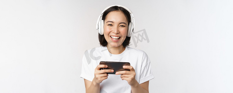 快乐的亚洲女孩玩家，玩手机，用智能手机观看，戴着耳机，站在白色背景上
