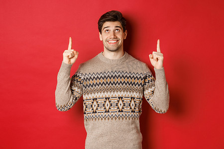 梦幻帅哥的肖像，想着圣诞假期，穿着冬季毛衣，指着并抬头看着新年促销优惠，站在红色背景上