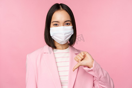 冠状病毒表现摄影照片_身穿医用面罩和西装的韩国女商人站在粉红色背景下，表现出大拇指朝下、不喜欢或不赞成的姿态