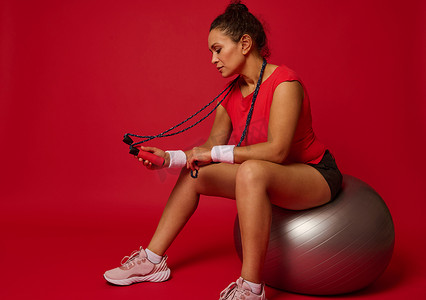 跳绳动态摄影照片_穿着运动服的漂亮健身女性坐在健身球上，手里拿着跳绳，在红色背景下锻炼后放松