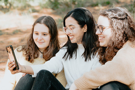 智能社区摄影照片_三个年轻女子在阳光明媚的日子里在森林里嘲笑她们正在看手机的东西、友谊和关怀概念