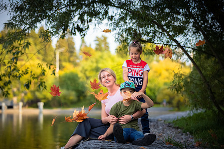 妈妈带着孩子坐在公园的池塘边，秋叶从上面飘落。
