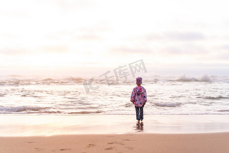 女孩望天空摄影照片_孤独的小女孩站在海滩上回到镜头，望着地平线。