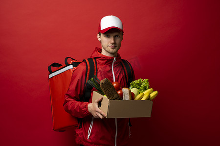 身穿红色制服的送货员拿着纸板纸盒，里面装着白色背景、工作室肖像中隔离的食物。