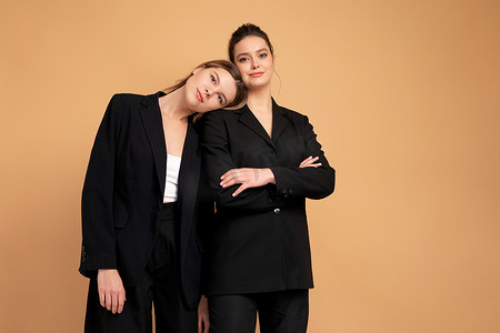 黑色西装女摄影照片_两名身穿黑色西装的年轻女商人站在工作室橙色背景