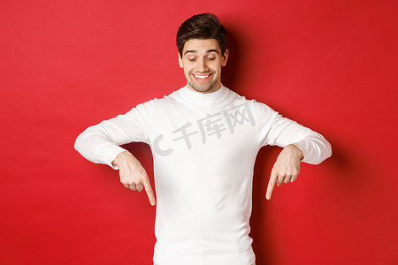 穿着白色毛衣的梦幻般的帅哥的形象，看着并用手指向下指着复制空间，关于新年和假期的广告，站在红色背景上
