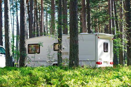 绿色拖车摄影照片_白色大篷车拖车停在松林的绿色草坪上。