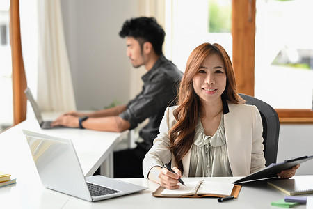 有魅力的亚洲女员工坐在公司办公室的笔记本电脑前，对着镜头微笑