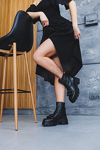 黑色高跟鞋图案摄影照片_没有高跟鞋的黑色皮靴，带扣，纤细的女性腿和灰色背景上的木椅。