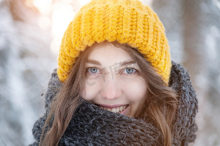 在白雪皑皑的森林背景下，一位戴着鲜艳羊毛帽子和围巾的微笑的年轻白人女子的特写肖像。